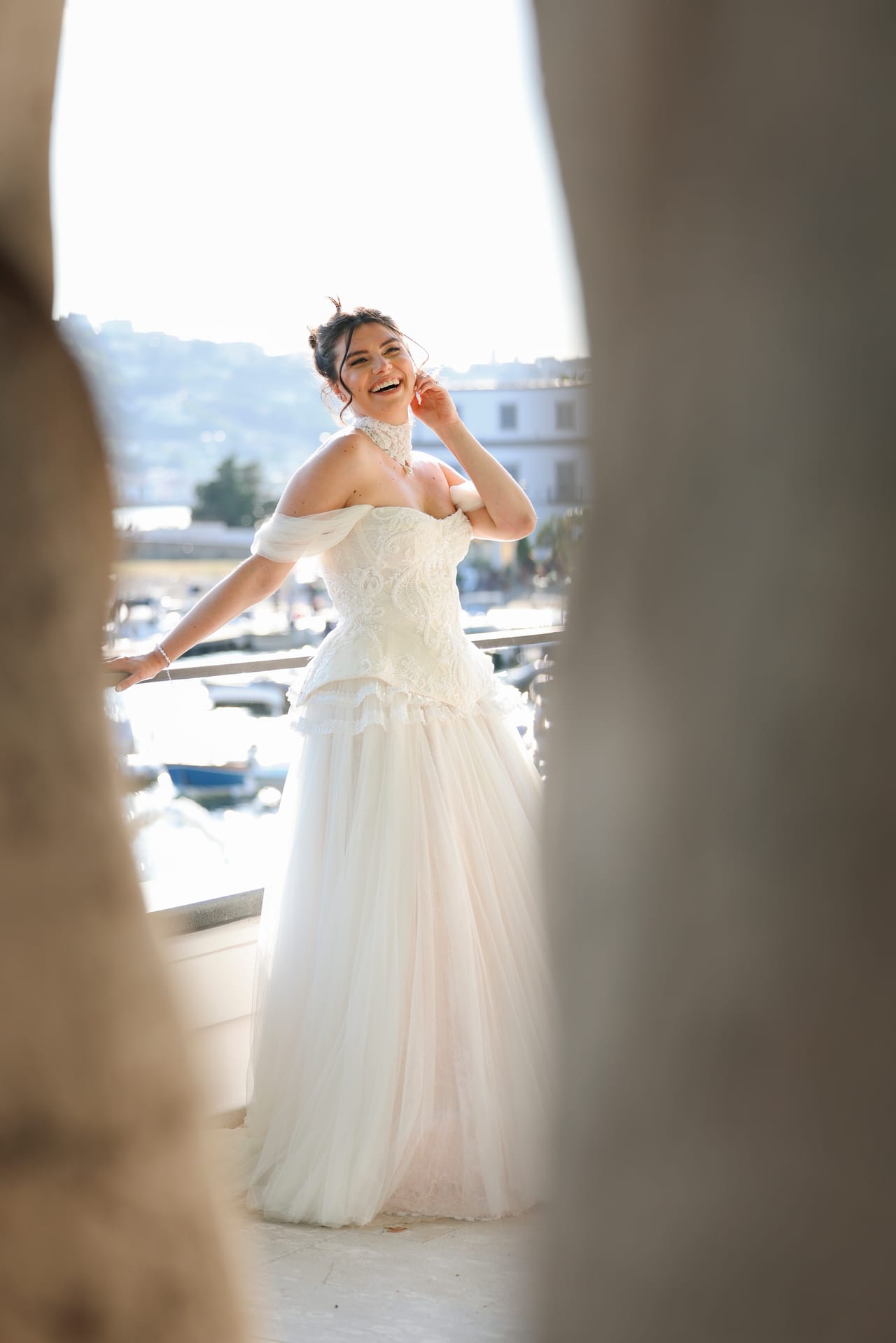 Sposa sorridente su un balcone affacciato sul porto, incorniciata da tende chiare