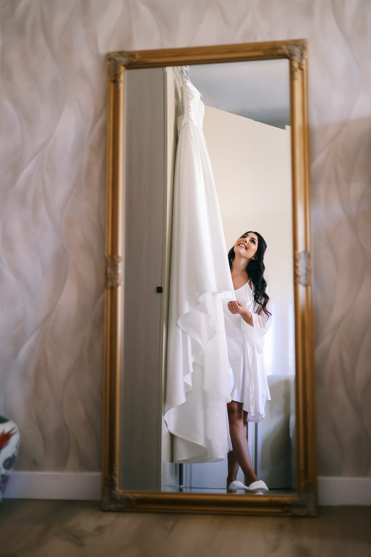 La sposa riflessa in un grande specchio dorato, accanto all'abito bianco appeso