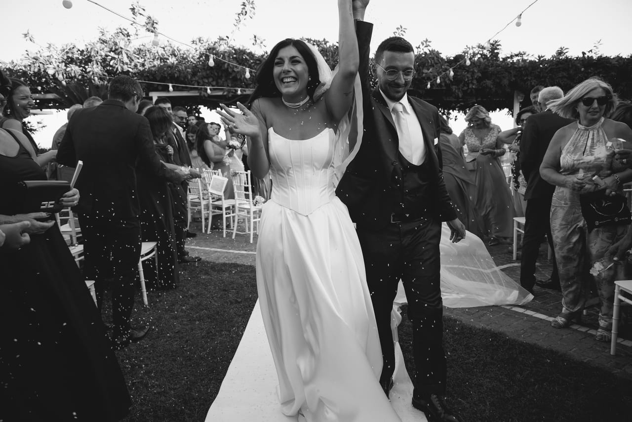 In bianco e nero, la sposa alza un braccio ridendo mentre attraversa gli invitati insieme allo sposo